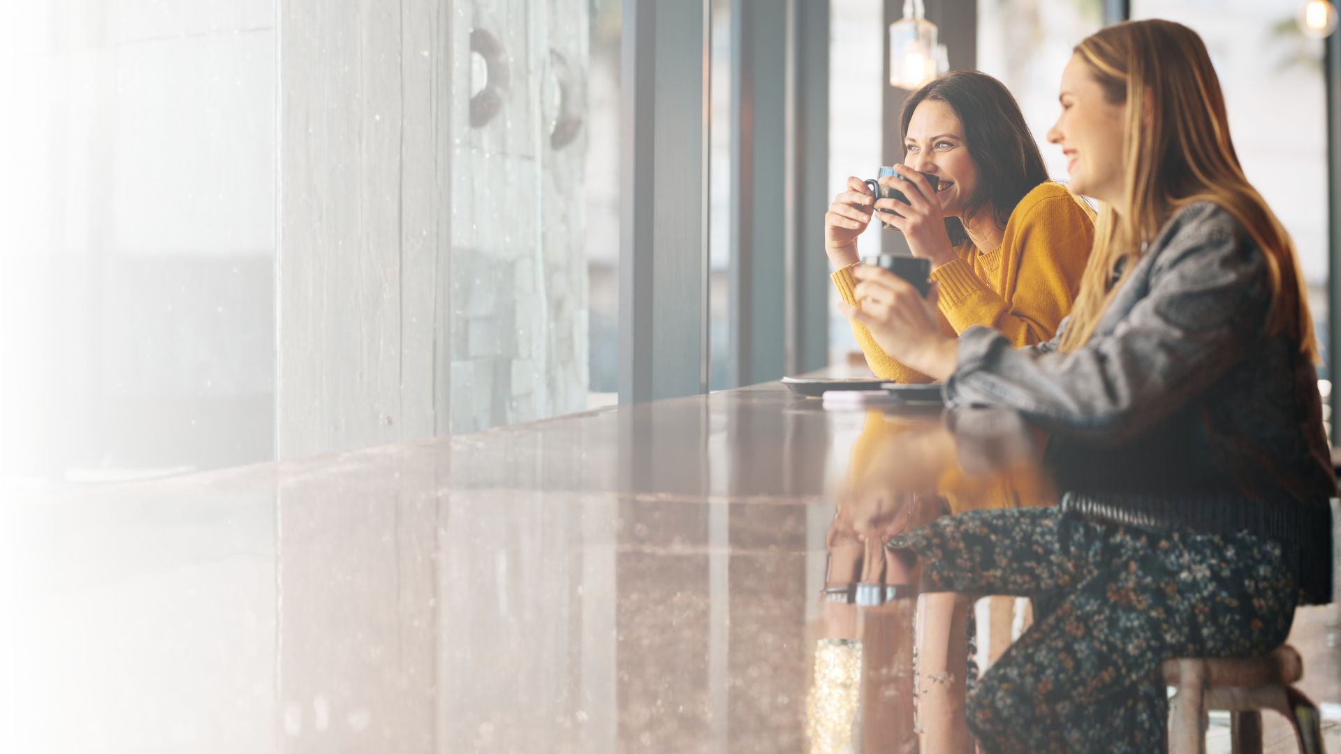 Twee vrouwen praten samen bij een raam met een kop koffie.